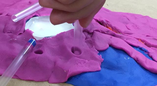 A straw is used to take a core sample from a play-dough volcano.