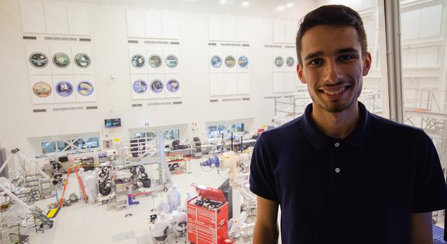 Adrien Dias-Ribiero poses for a photo in the gallery above the clean room at JPL with the Mars 2020 rover behind him.