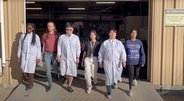 Six students of various ethnicities and genders walk out of a garage housing a model of the Perseverance Mars rover.