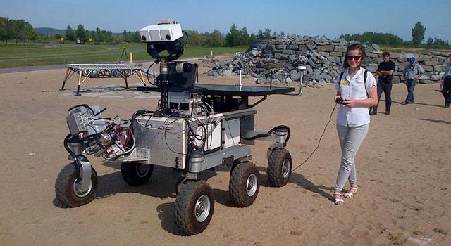 Brittney Cooper stands in a sandy area holding a controller attached to a rover