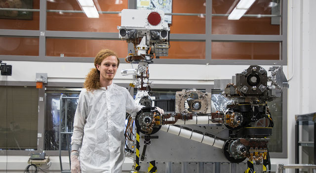 Jeff Carlson stands in an open room posing next to an engineering model of the mast for the Mars 2020 rover