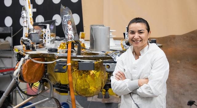 Marleen Sundgaard stands on gravel in a tall room with a test version of the InSight Mars lander behind her.