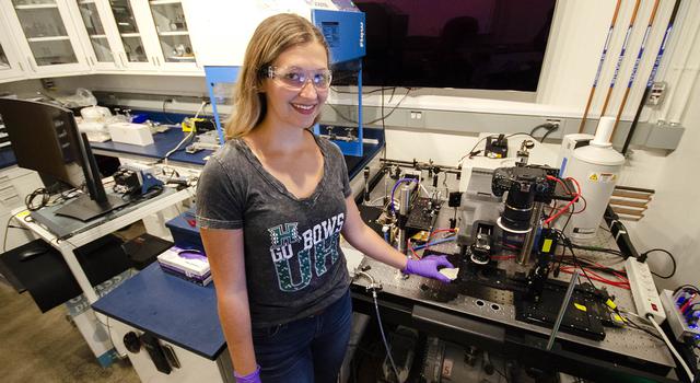 Schelin Ireland, wearing lab goggles and purple gloves, holds a sample under a laboratory version of the SHERLOC instrument for Mars 2020.