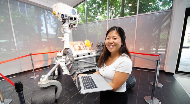 Vivian Li holds a computer and poses for a photo in front of a full-size model of the Mars rover Curiosity.