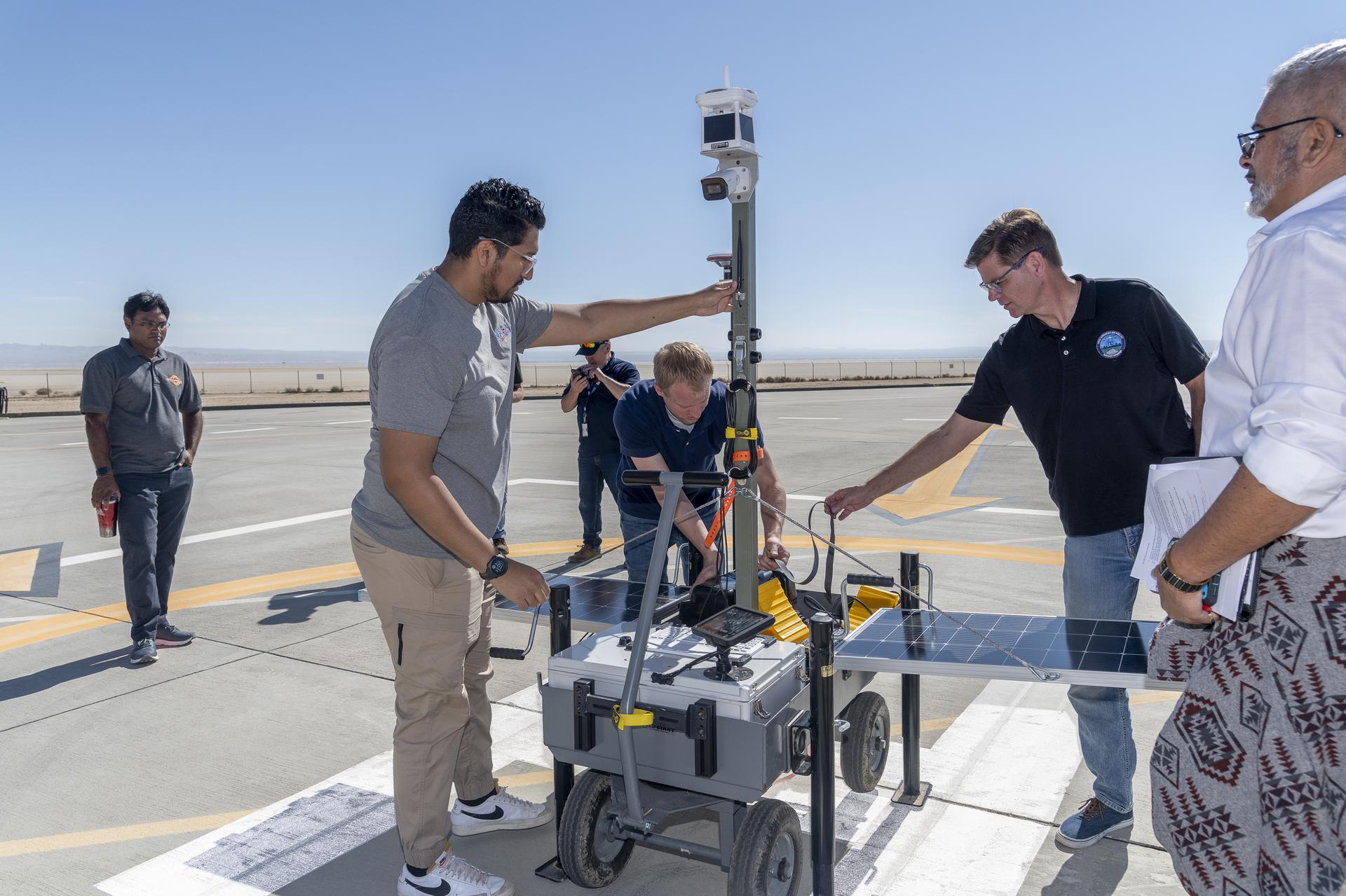 A man wearing clear glasses is standing to the left of the image wearing a gray shirt and tan pants. His right arm is touching a cart holding technical instruments including two solar panels sticking out from each side. There are several other people surrounding him.