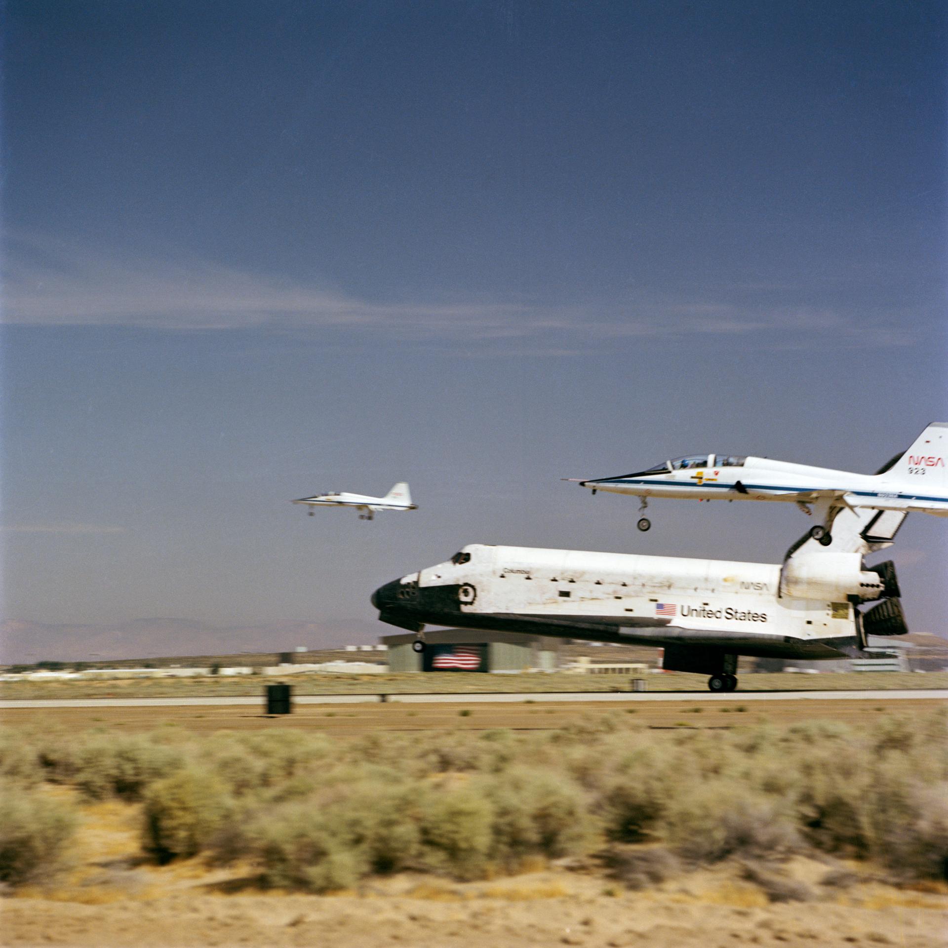 Space Shuttle Columbia lands at Edwards air force base with two chase planes flanking it.