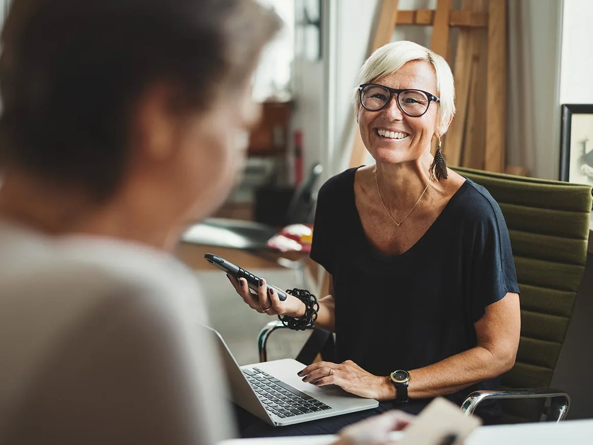 Woman with a laptop and phone smiling at a colleague