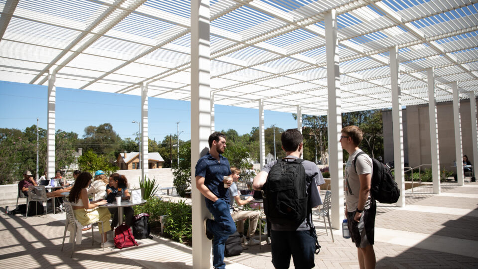 A student in a blue tee shirt and jeans stands and talk to a student wearing a backpack and dark pants and a students wearing a backpack, gray tee shirt, and blue shorts.