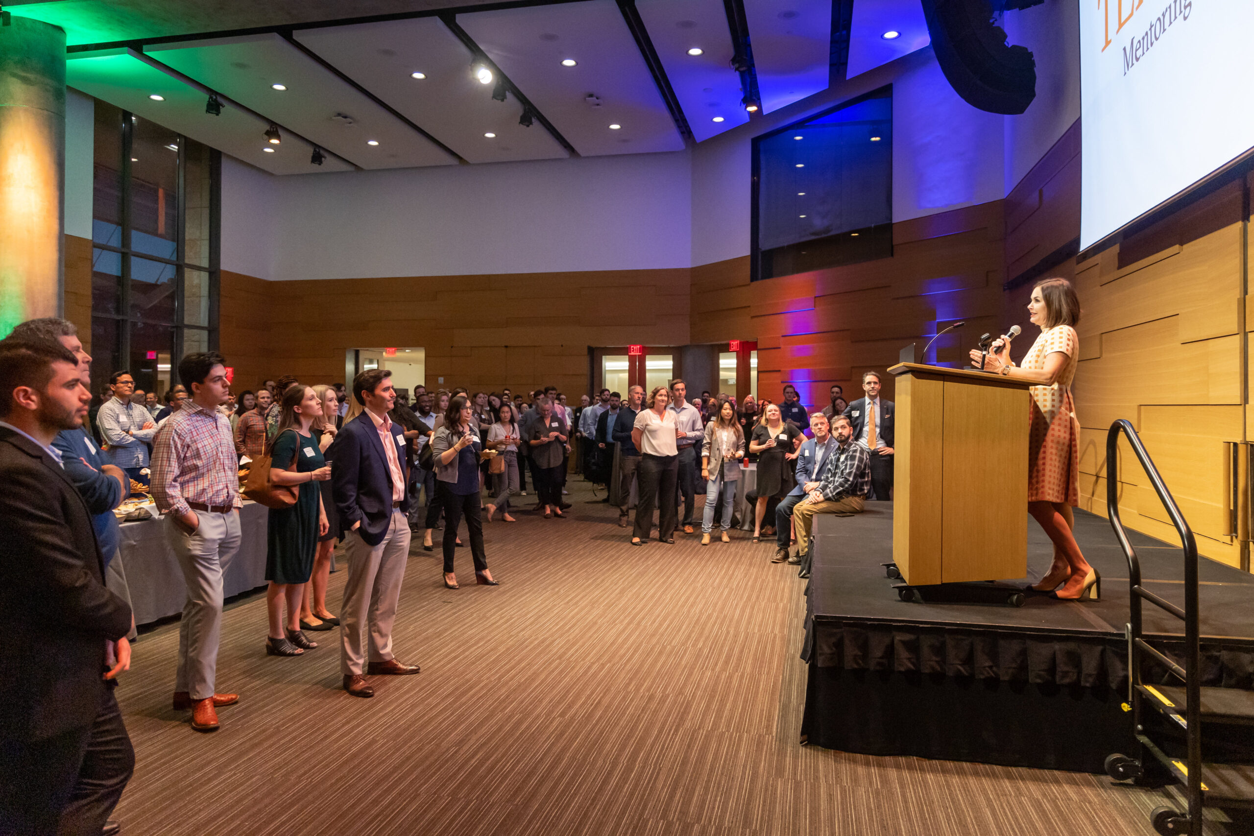 Mentors and mentees stand in front of stage listening to Rémi Ratliff speak at podium