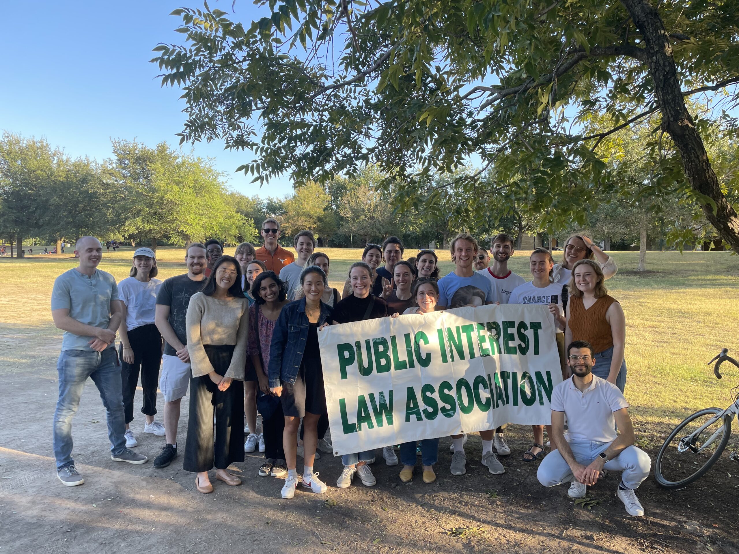 A group of 25 students stand outside under a tree holding up a banner that reads Public Interest Law Association