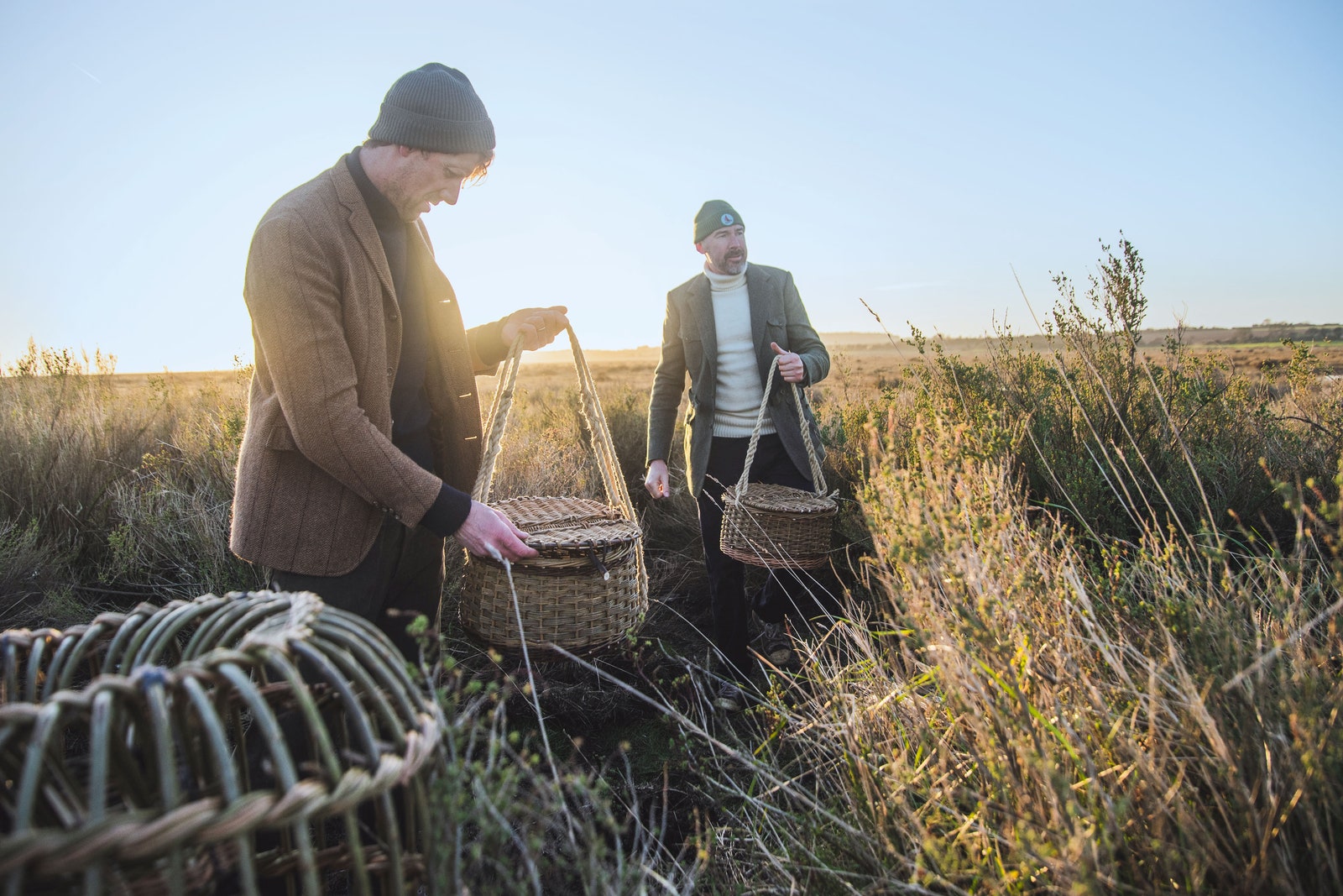 Two men dressed in jackets and hats holding baskets in a grassy field