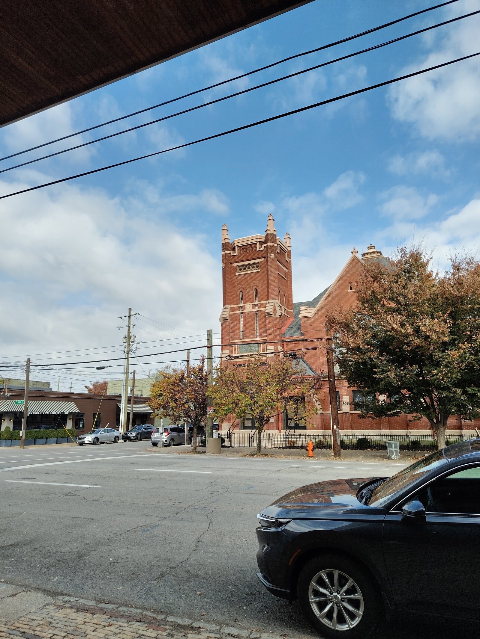 View from across the street of a brick building with a tower and parked cars along the sidewalks