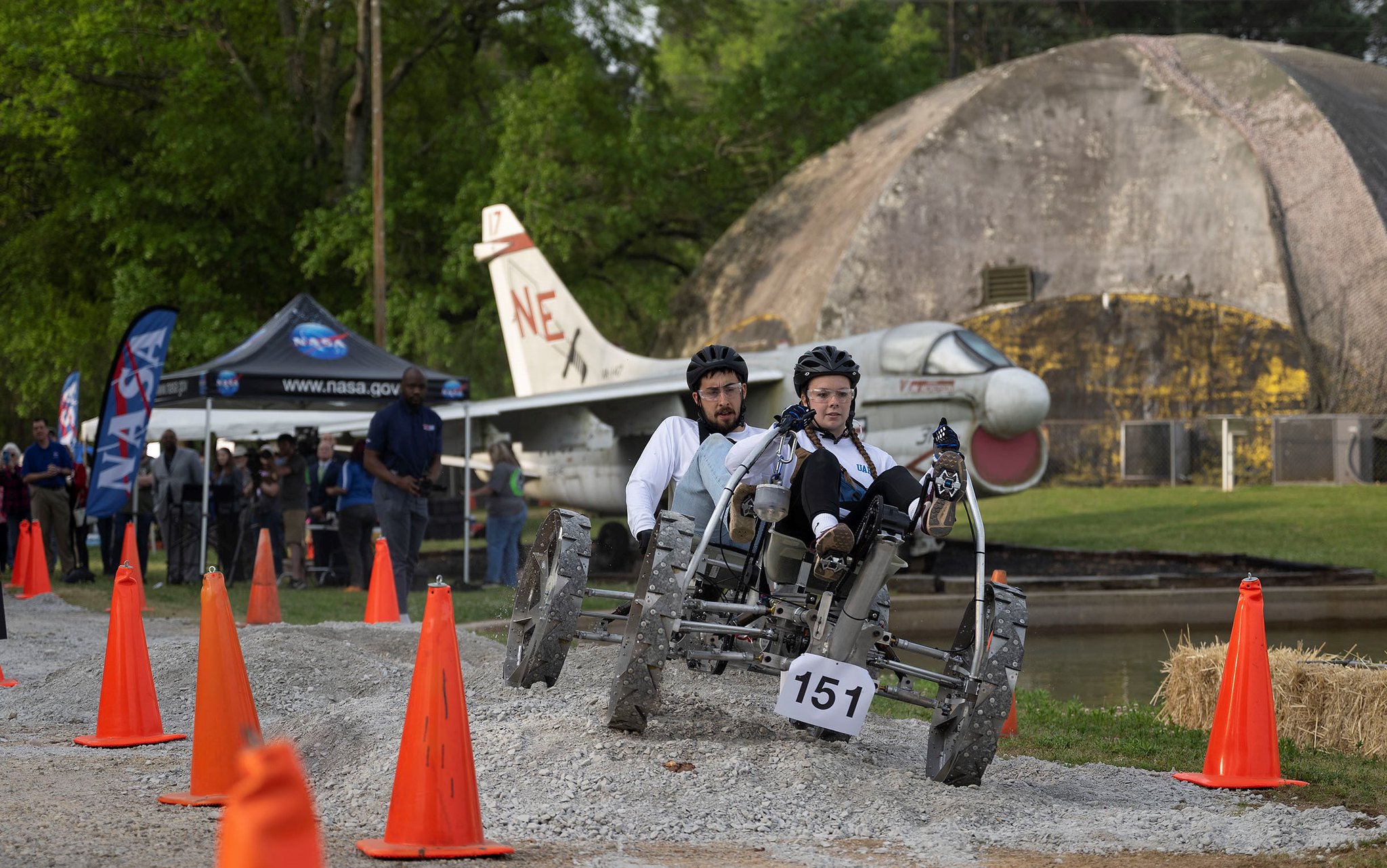 Two Human Exploration Rover Challenge competitors pilot their human-powered rover through the course at NASA's Marshall Space Flight Center. The duo wear bicycle helmets and pedal their metallic rover over gravelly terrain. Throughout the foreground, large orange cones guide the rover's path. In the background, onlookers watch from beneath a large NASA pop-up tent. A large rock and stationary jet sit in the far background. Credit: NASA