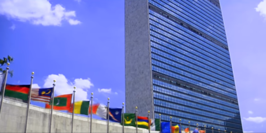 Flags representing multiple countries wave in the wind outside a tall building.