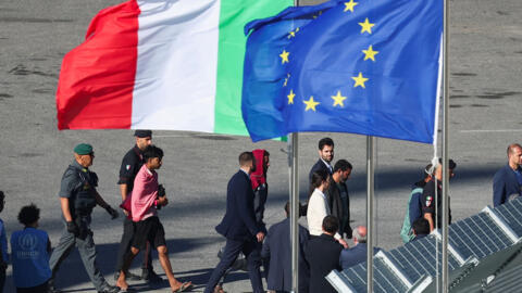 Migrants and security officials walk next to the European Union flag and Italian flag as they disembark from the Italian navy ship Libra that arrived in Albania as part of a deal with Italy to process thousands of asylum-seekers caught near Italian waters, in Shengjin, Albania, 16 October, 2024. 