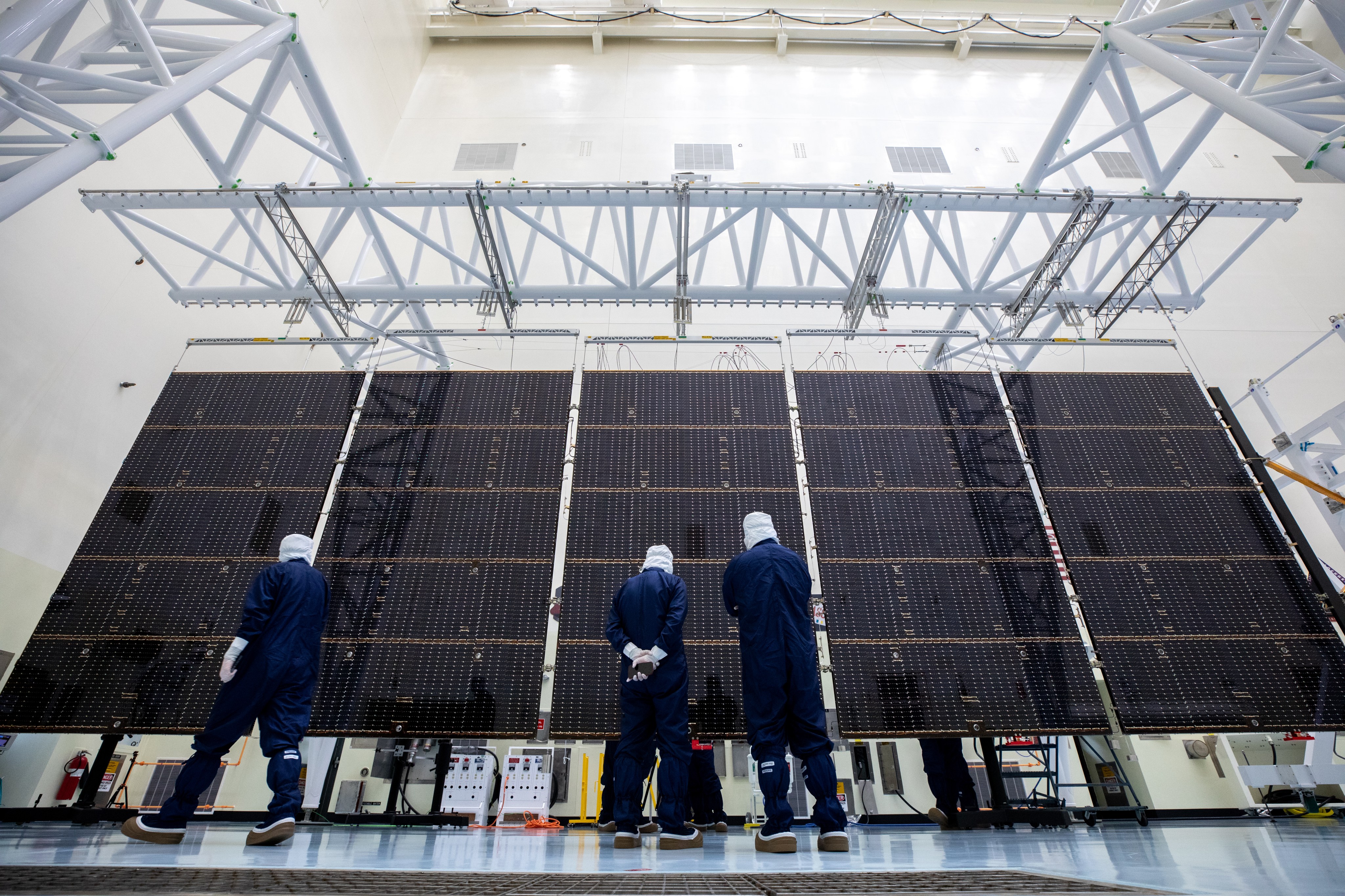 Three workers in blue protective jumpsuits with white head covers stand in front of part of the solar arrays for Europa Clipper.