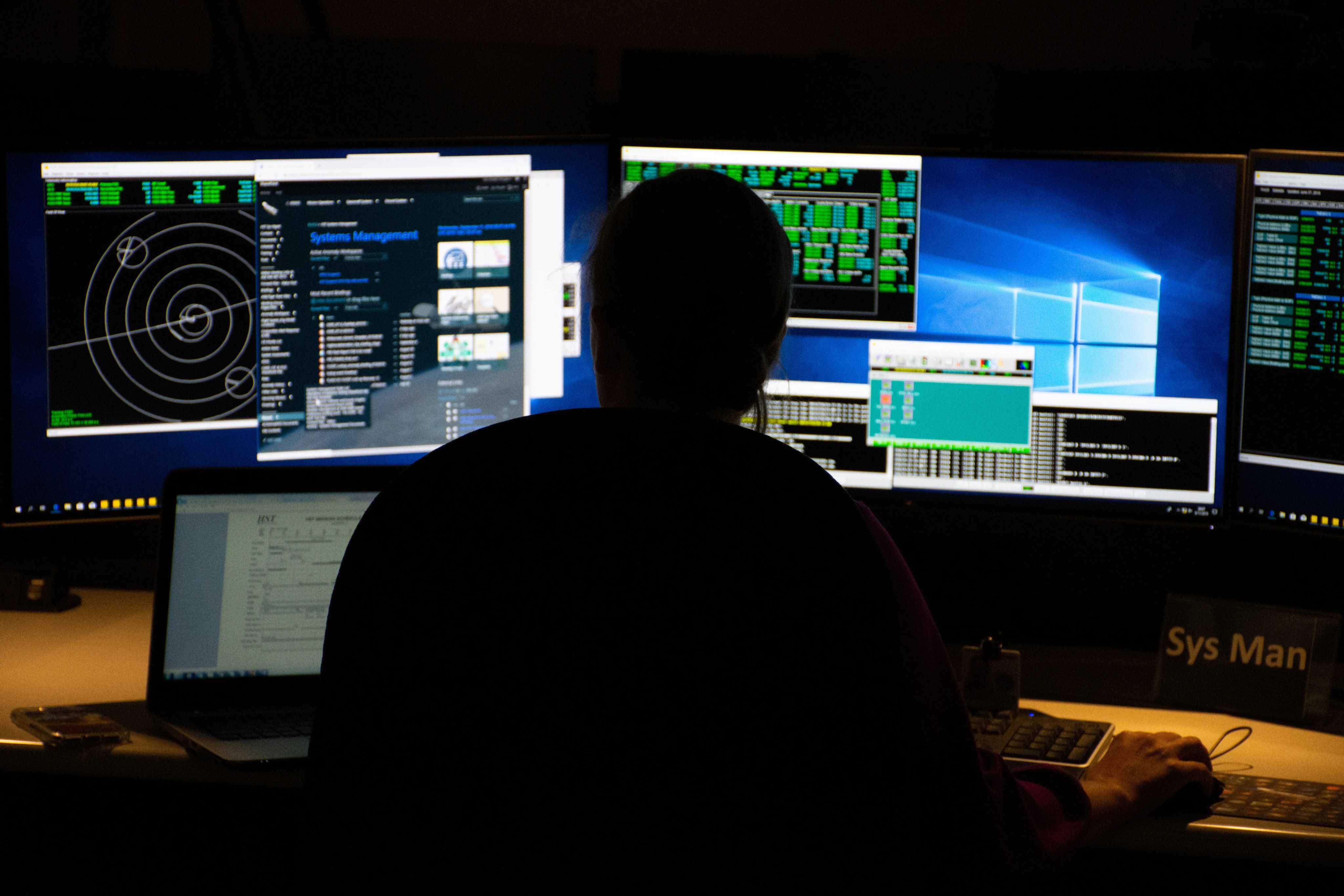 A person sits in dim lighting with their back to the camera, facing two computer monitors displaying data from Hubble.