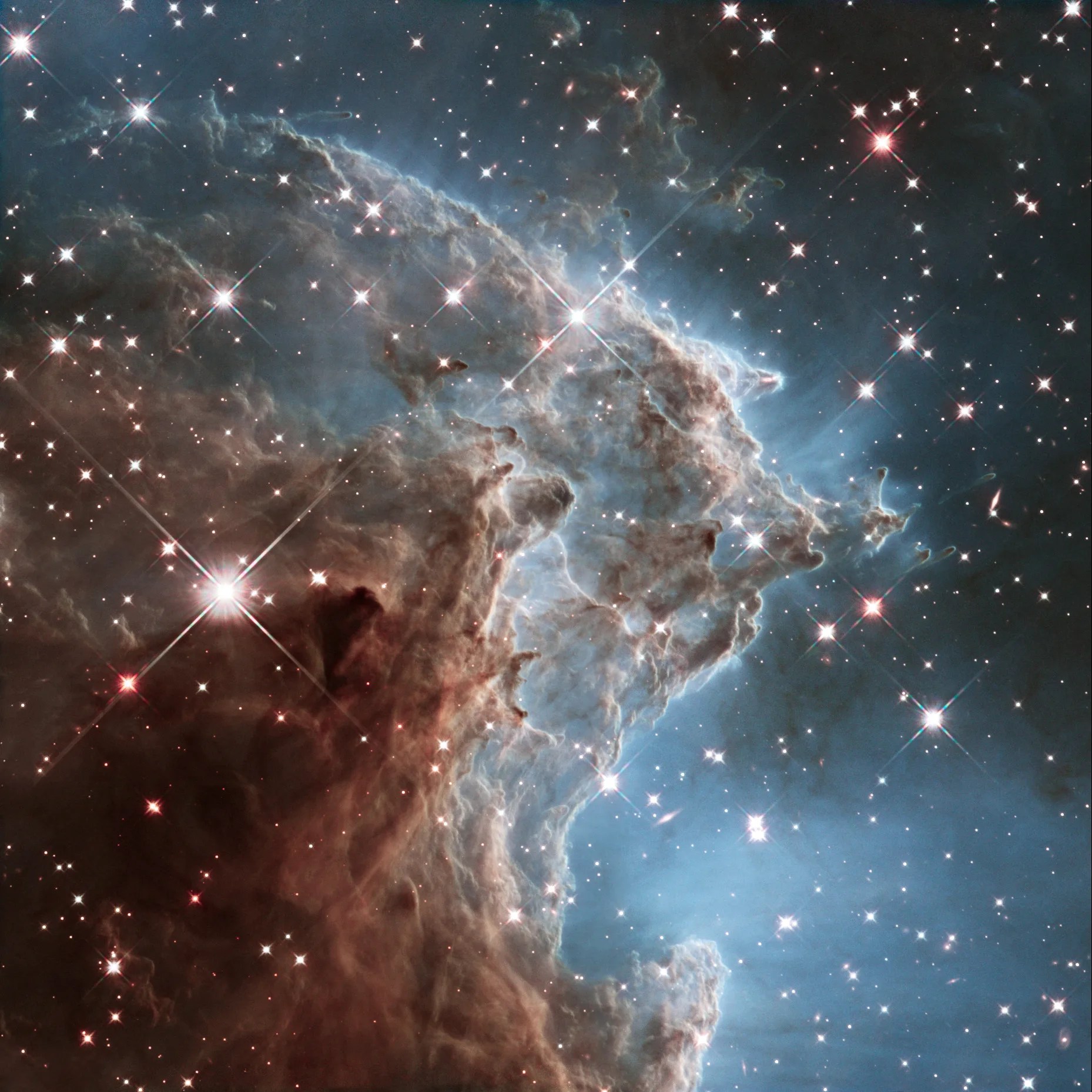 Lower -left quadrant is filled with rusty-brown dust cloud dotted with a few bright stars. Hazy pillars of gas and dust rise up toward the right. A bluish-white glow fills the lower-right quadrant. The haze is more transparent as you move toward the upper right corner. Background is dotted with stars.