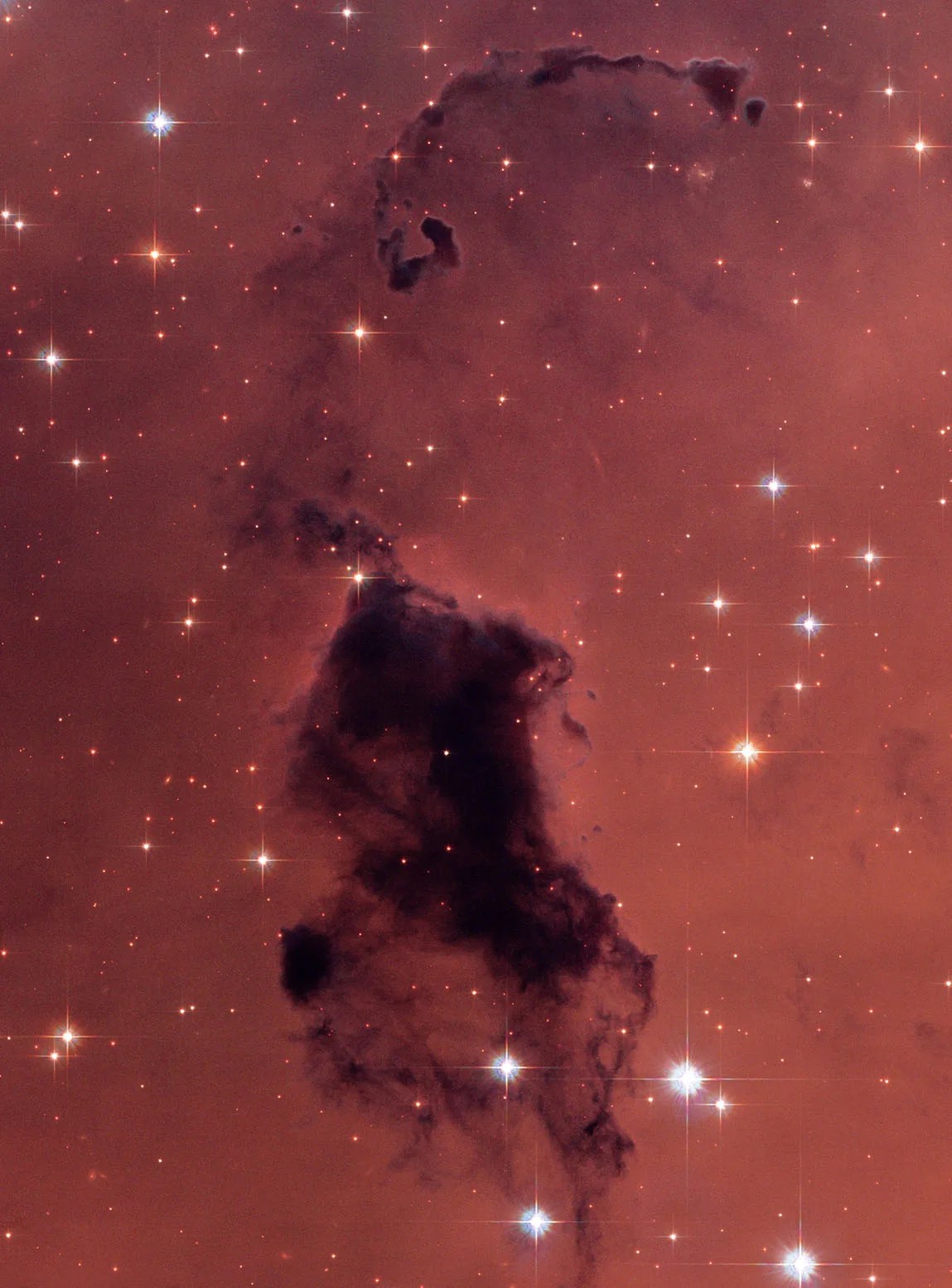 Glowing reddish gas is speckled with bright-white stars. In the foreground is a dark, vertically extended cloud of gas and dust that blocks the light of objects behind it. The largest part of the dark cloud (Bok Globule) extends from image center to bottom. Tenuous clouds extend vertically above the main globule toward the image top.