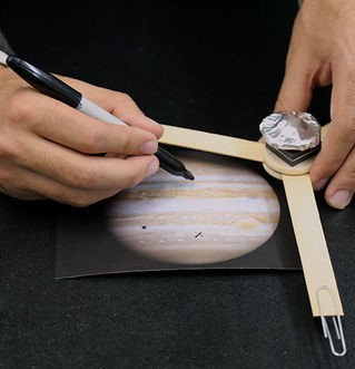 Closeup of hands making a Juno model out of popsicle sticks and foil connected in a 3-pronged model