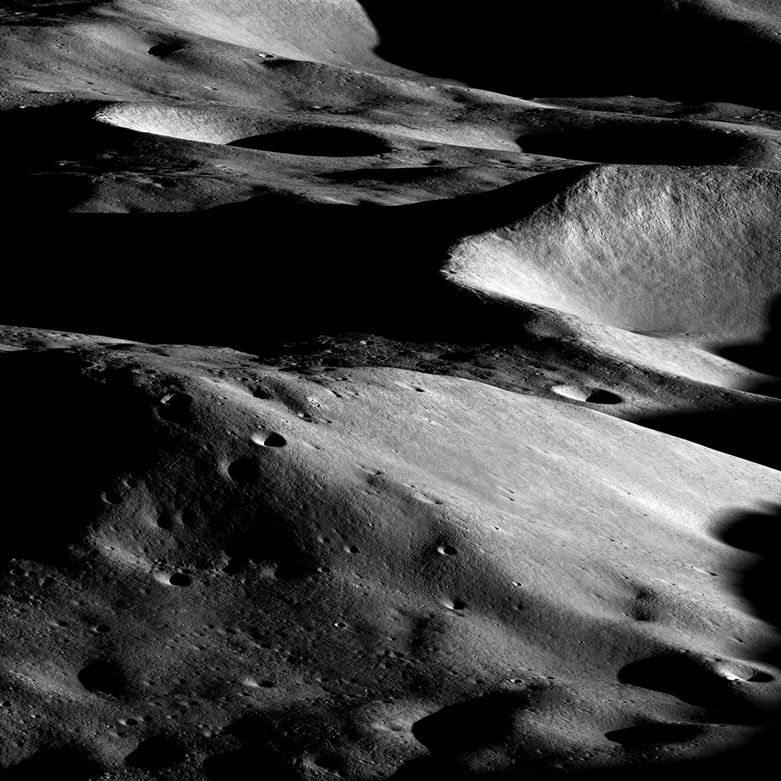 Dramatic grayscale image of a mountainous lunar landscape. Large craters with black shadows inside are visible in the background. In the foreground, a mountain is sunlit from the right and pocked with small craters.