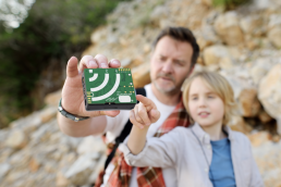 A man and a child examine an AudioMoth recorder, a green square-shaped device about the size of a deck of cards.