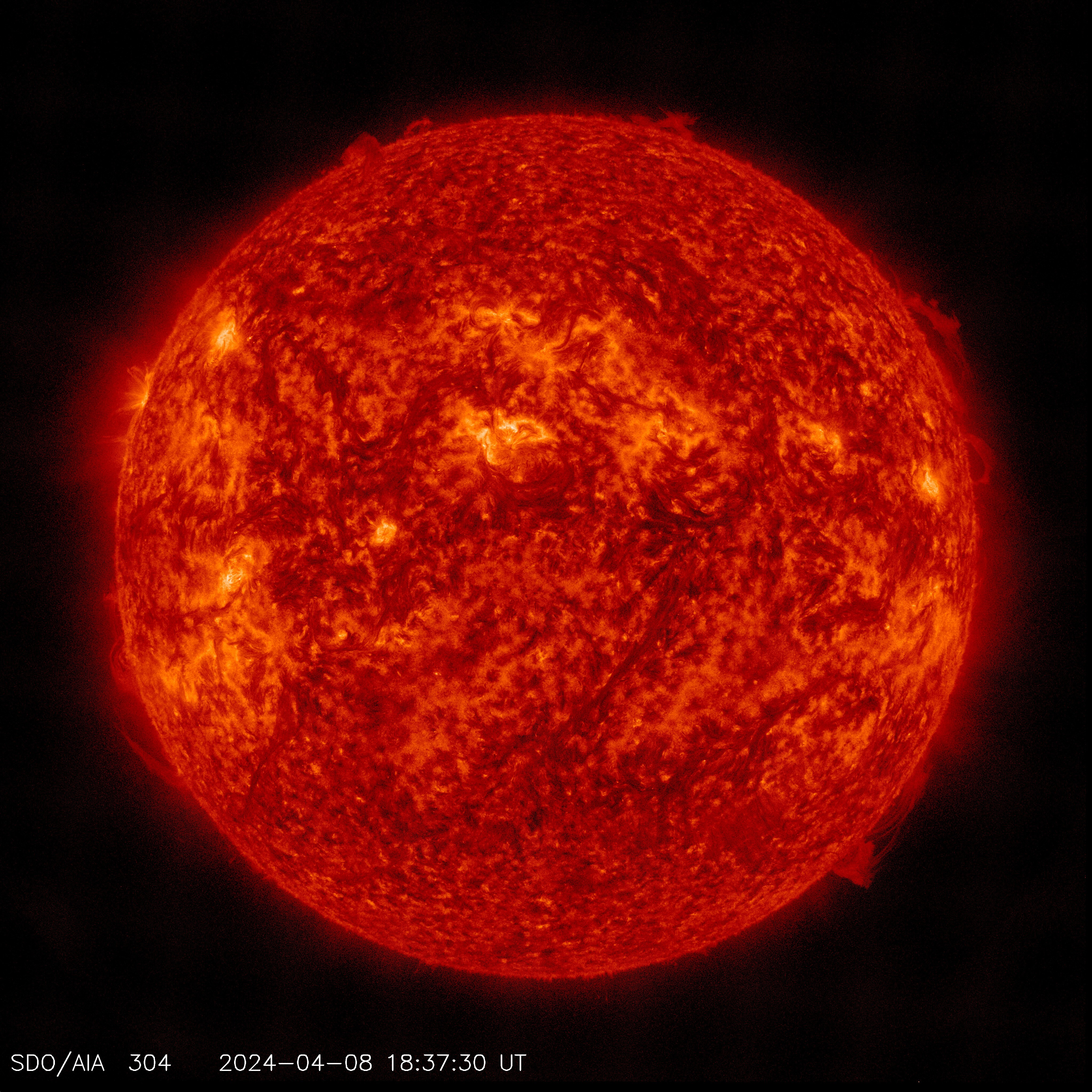 Against a black background, the Sun seen in red, with spots of orange and black swirling. On the bottom right area of the star, there are spurts of solar material seen against the black background.