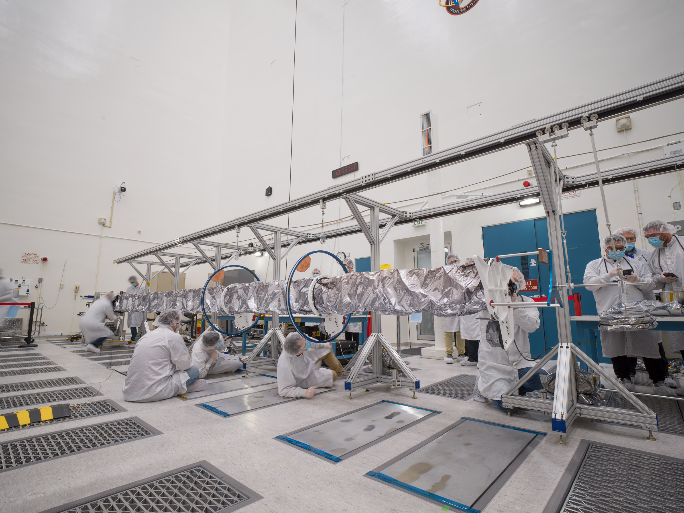 Workers in white protective clothing sit, and crouch on the floor as they unfurl a long, shiny silver-colored piece of equipment. Round hoops and aluminum-colored tripods help support the equipment.