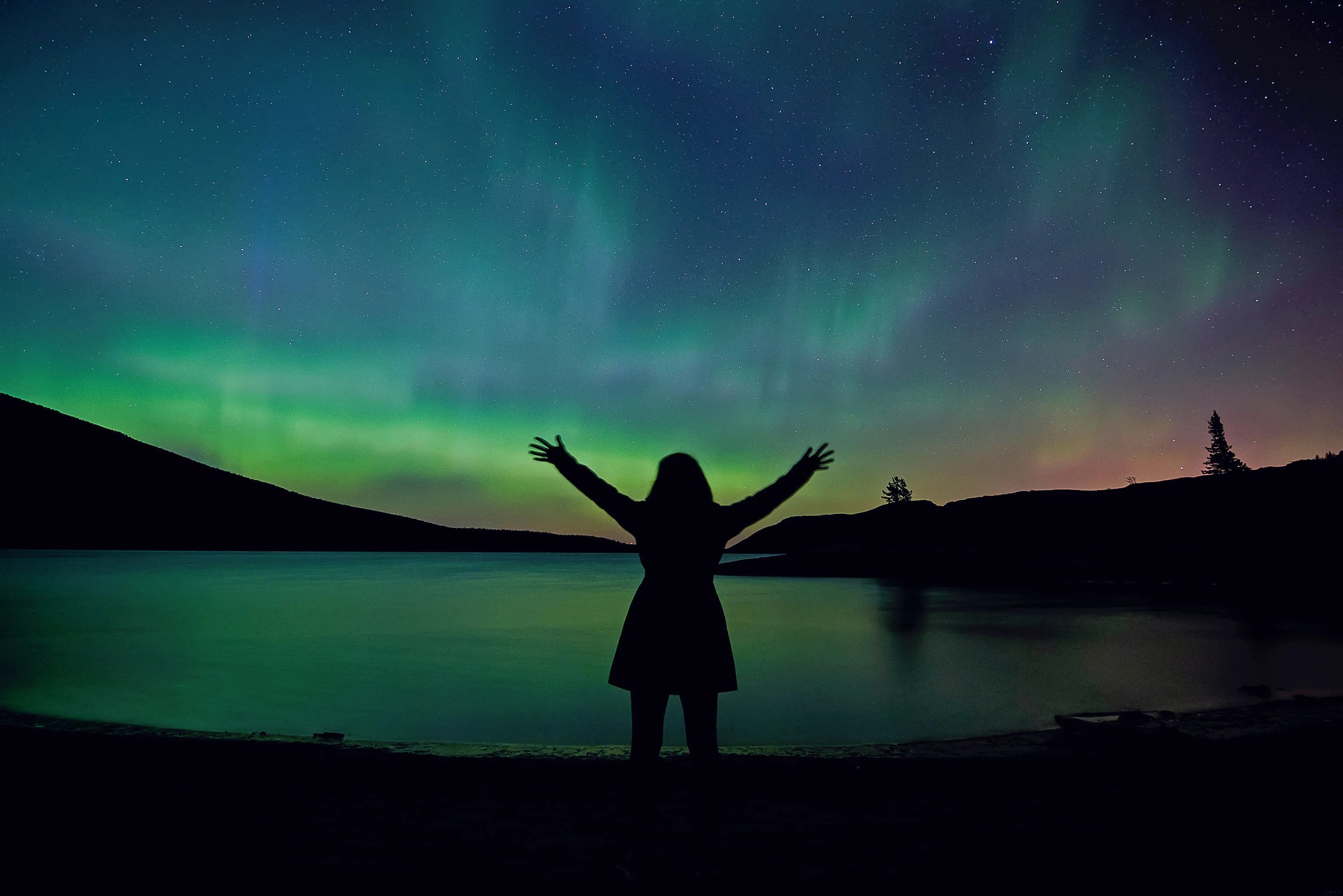 A woman with outstretched hands is silhouetted against a sky glowing with colorful auroras.