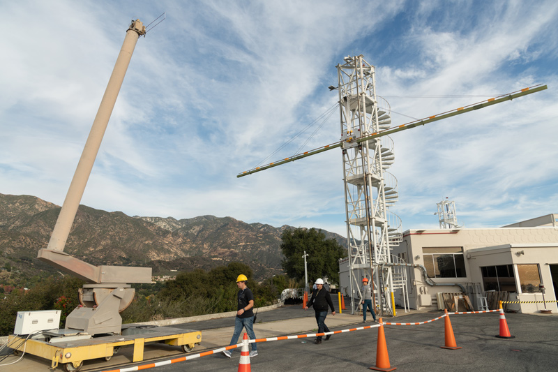 Three people work on a Europa Clipper instrument in a ooned-off area in a hilly part of JPL. Equipment includes a white tower with a spiral staircase to reach the instrument - which looks like a long white board with orange markings. The tower is next to a beige-colored building. A beige-colored lift sits nearby on a yellow platform.