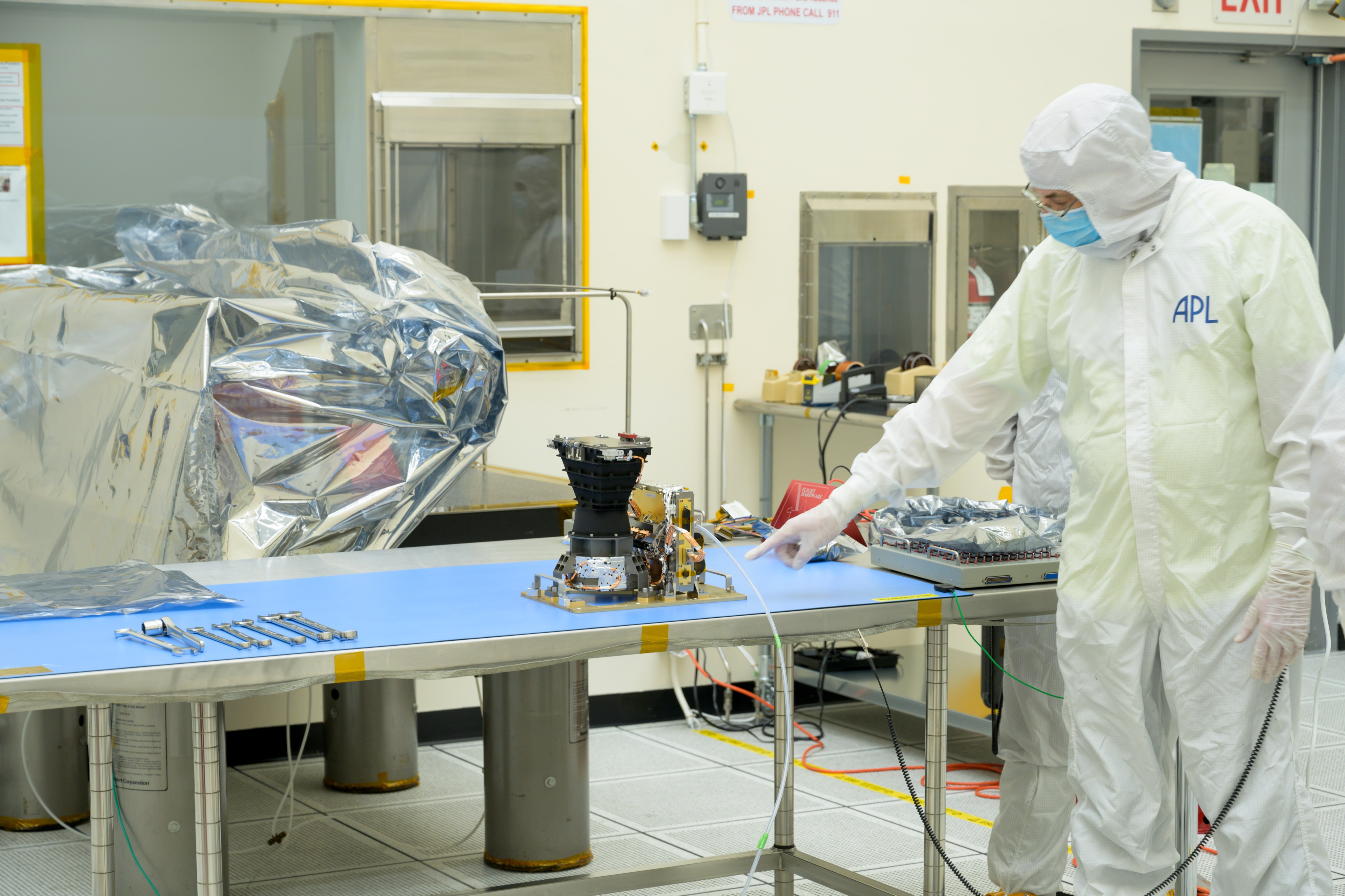 A small dark gray instrument that widens at the top sits on metal table with a blue cloth and tools on it. A worker in white protective clothing with the initials APL on the left side points at the instrument.