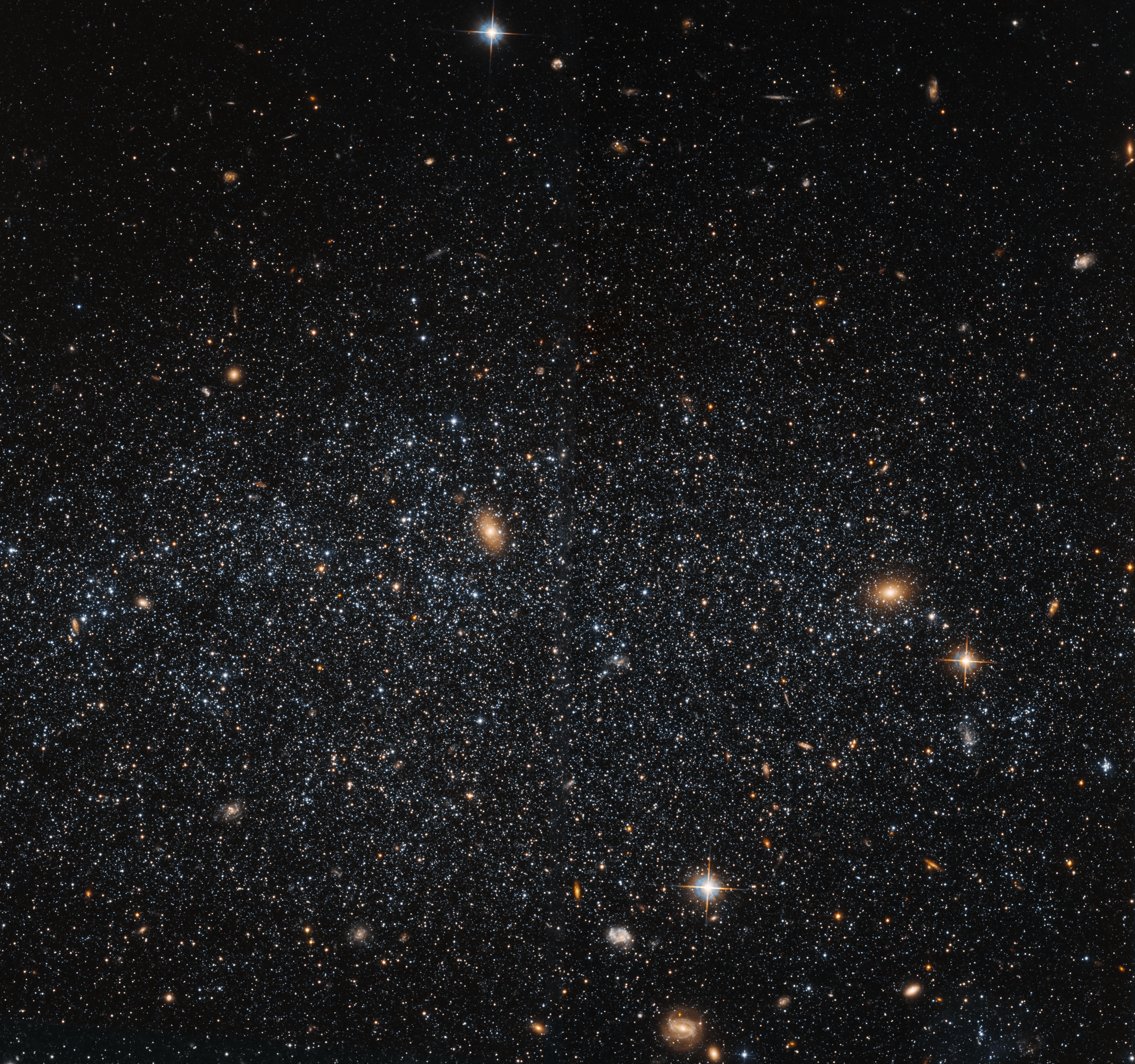 A black background dotted with stars and distant galaxies. The distribution of stars increases toward image center, forming the loose oval shape of the irregular galaxy Leo A that stretches horizontally across the image. Three foreground stars shine brightly – one located at center-top, and two others in the lower-right quadrant of the image. Each holds four bright barbs (called diffraction spikes) that extend outward from the star.