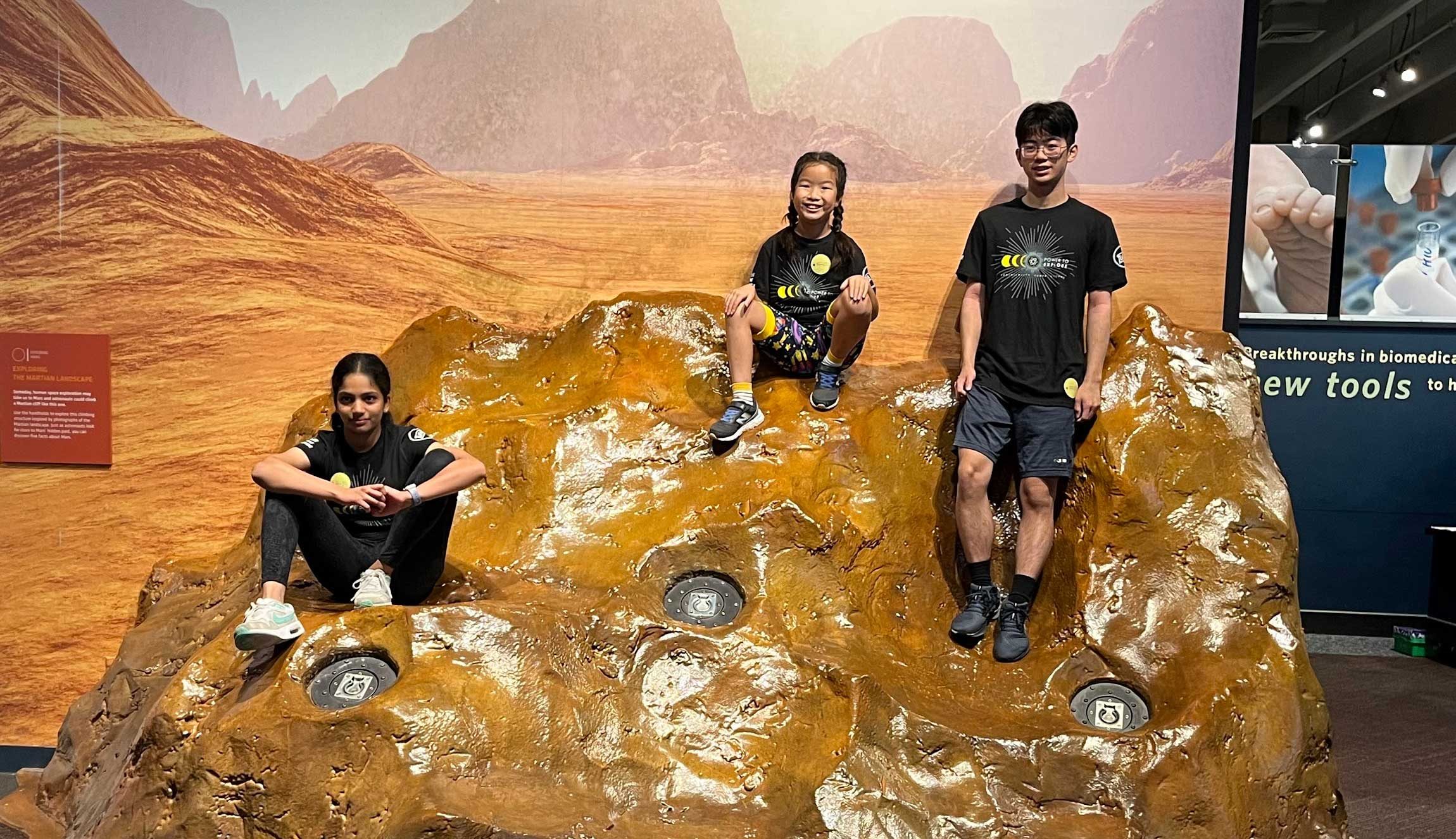 Thre children, a boy and two girls, sit on a model rock formation.