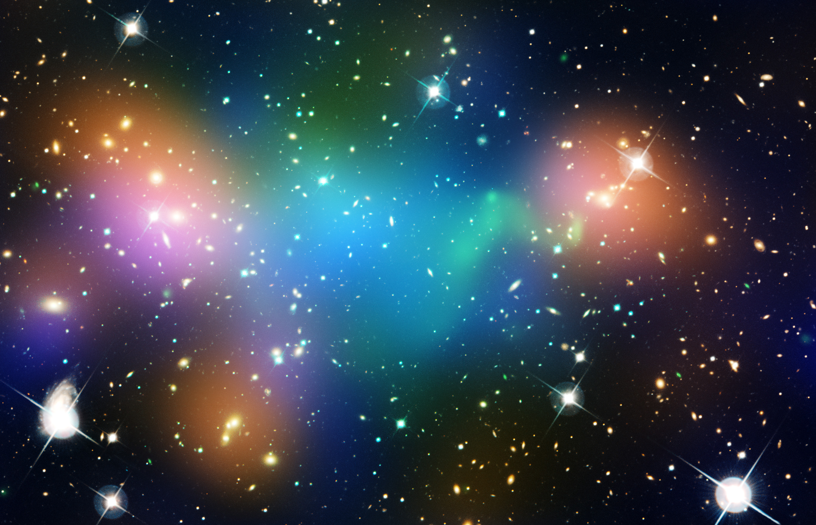 Green, orange, blue and purple blobs of glowing light mingle with the shining galaxies in a massive cluster. Blue and green is concentrated in the middle and orange and purple is in splotches on the outskirts.