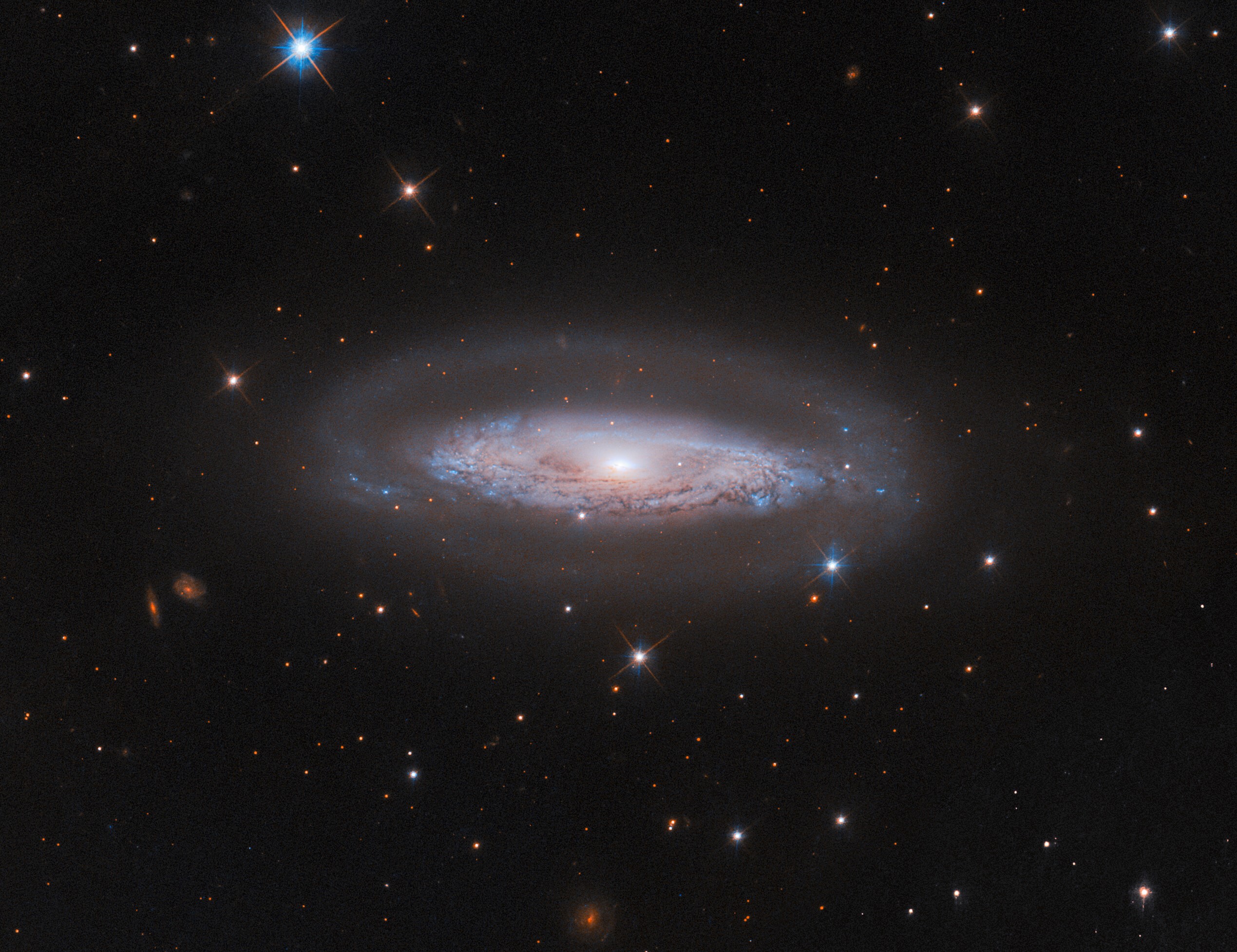 A spiral galaxy with a bright-white core, a glowing disk thick with swirling patterns of dark dust, and a faint halo around the disk. It is on a black background with a few small, distant galaxies and some foreground stars around it.