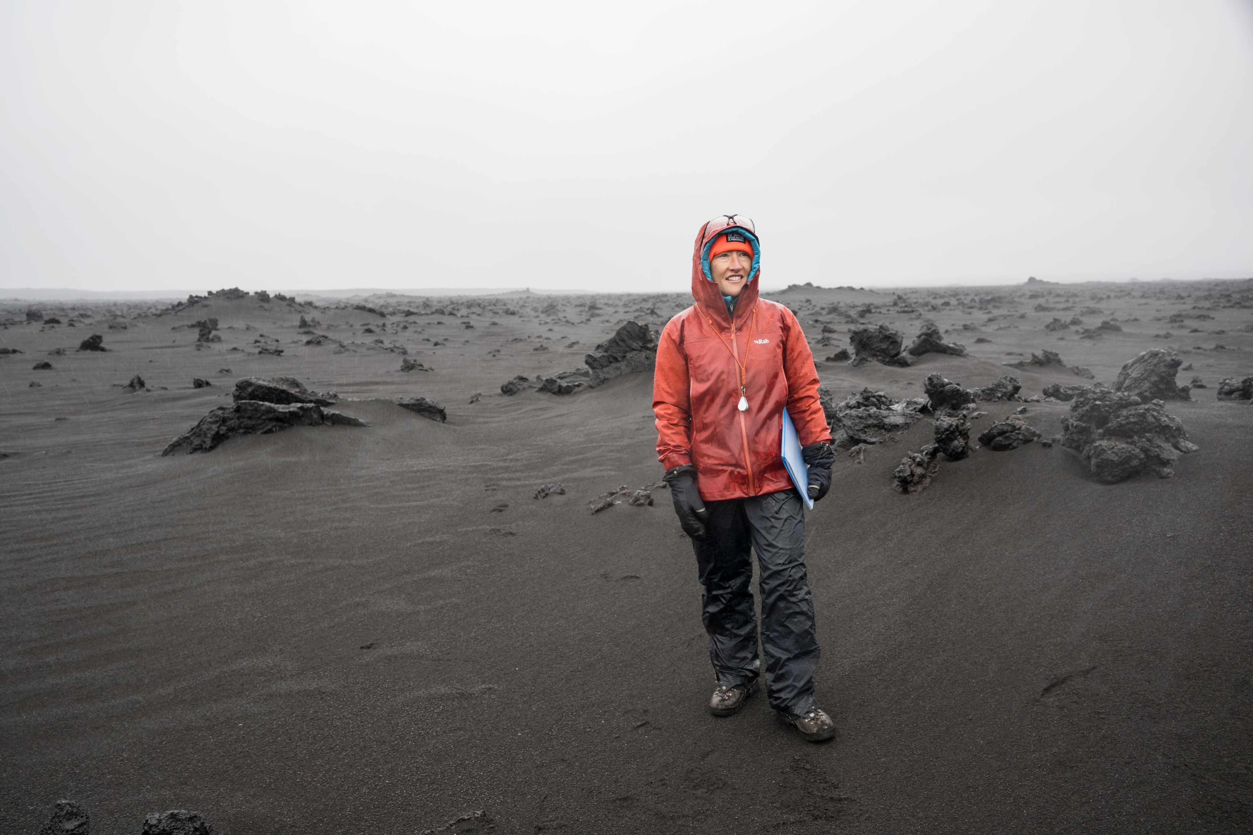 On a stark, barren landscape of dark grey rocks and dust, a smiling person dressed in rain gear and warm layers holds a clipboard under her arm.