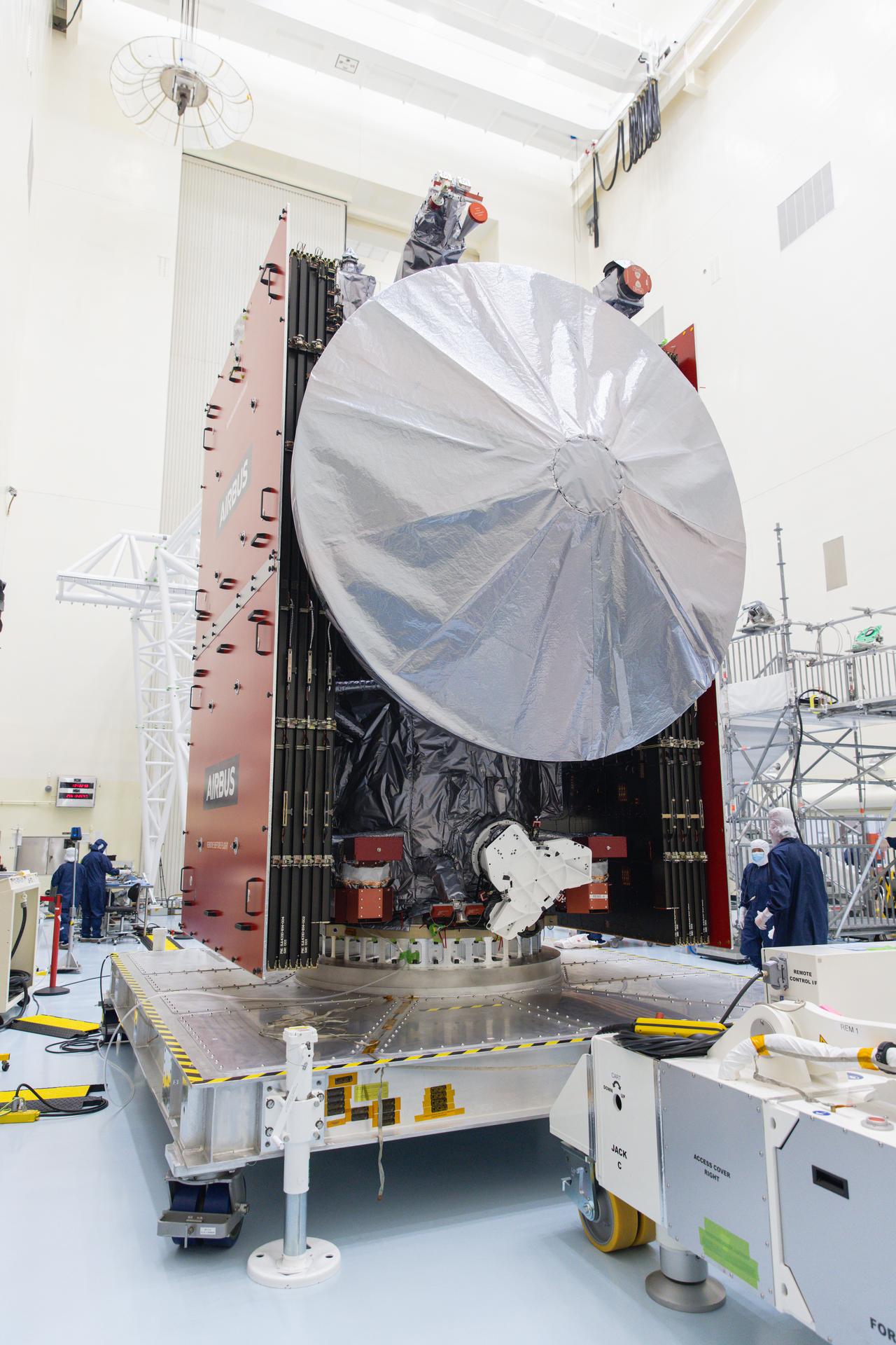 Europa Clipper sits on a silver work stand. The spacecraft's high gain antenna is in the front and center of the image. The folded solar panels are attached to each side of the spacecraft. At least four workers in blue jumpsuits and white head covers are to the sides of the spacecraft.