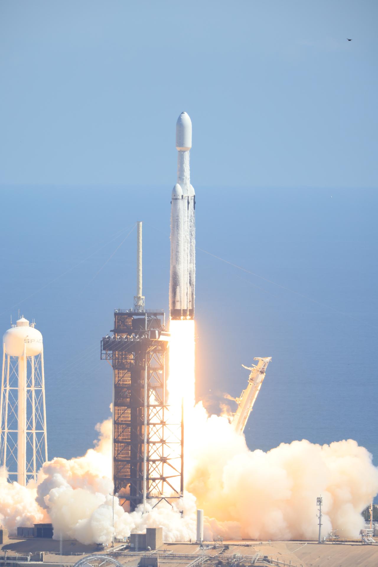 A white rocket and side boosters with a spacecraft enclosed in a protective fairing are shown just above the launch pad. White fire shoots out of the bottom of the rocket while billowy white smoke covers much of the launch pad.