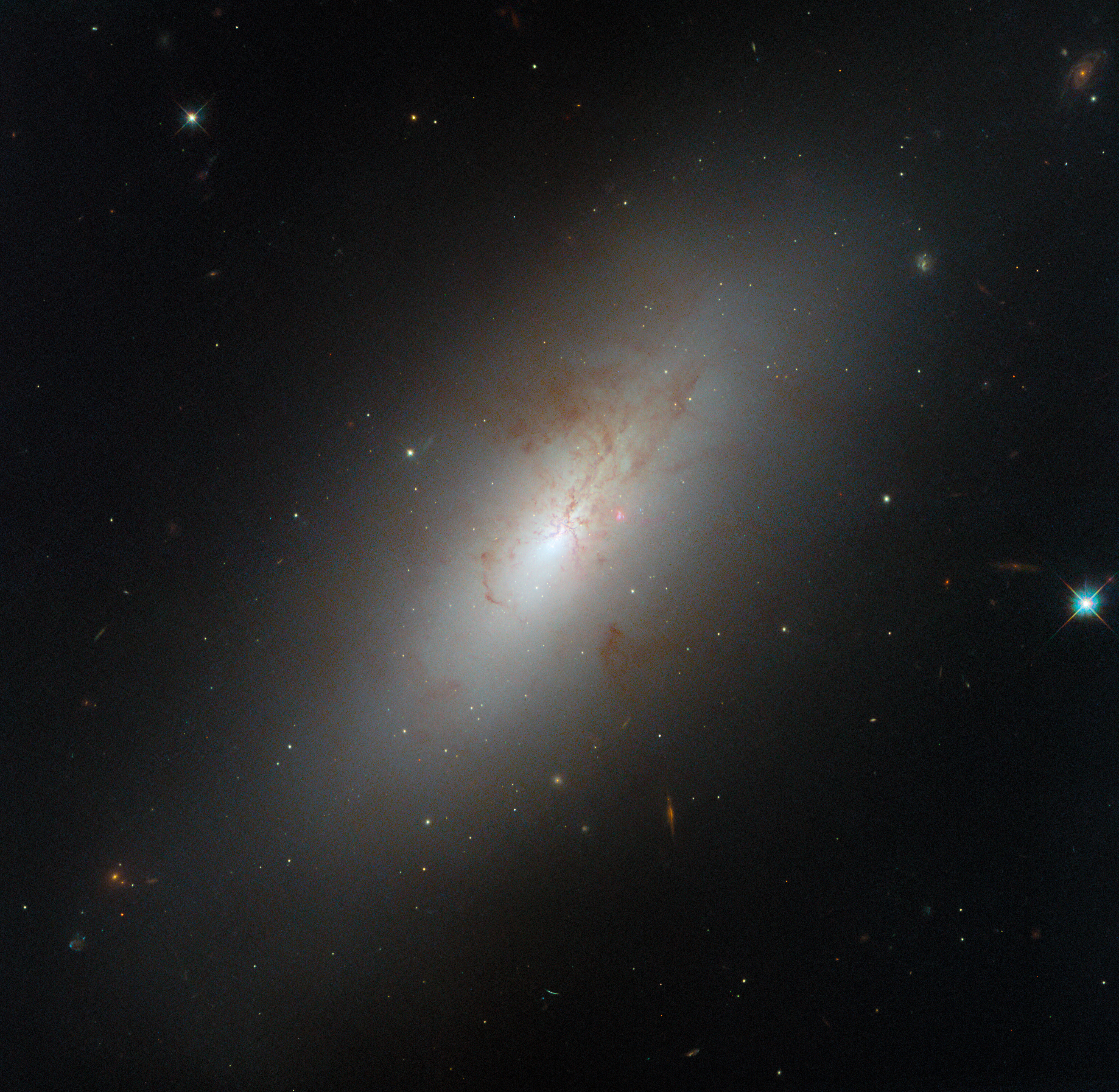 An oval-shaped galaxy seen tilted at an angle. It glows brightly at its core and radiates outward, dimming toward the edge of the oval. Reddish-brown, patchy dust spreads out from the core and covers much of the galaxy’s top half, as well as the outer edge, obscuring some of its light. Stars are visible around and in front of the galaxy.