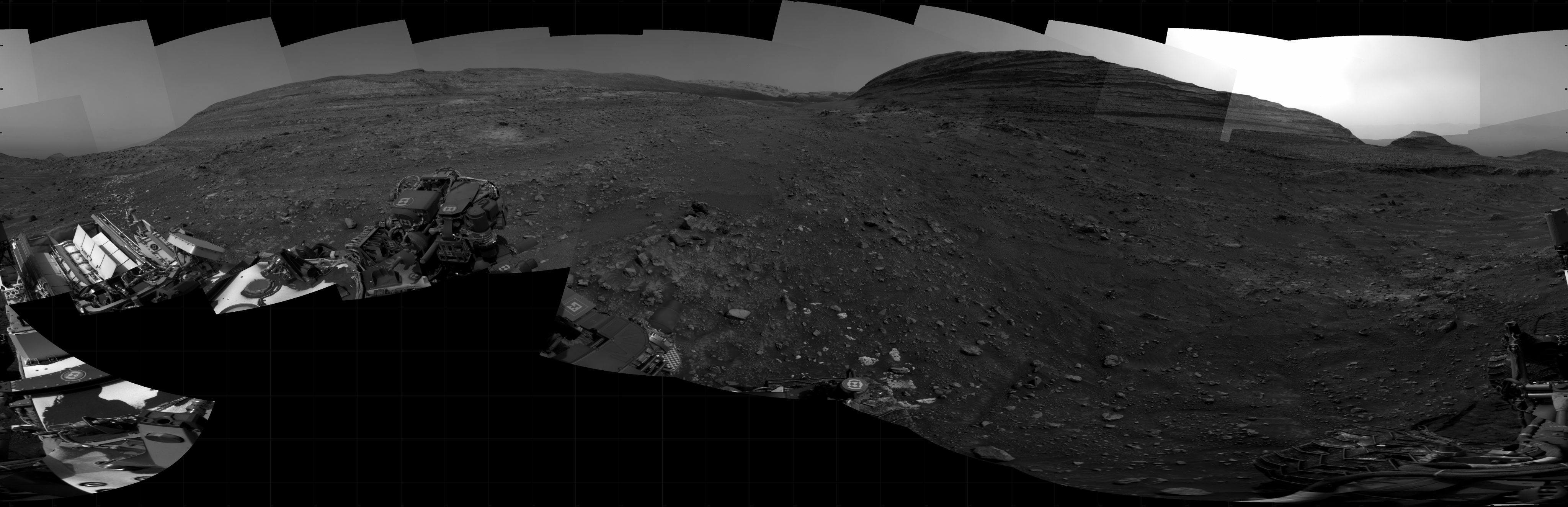 A grayscale panoramic image of the Martian surface shows both sky and ground with a hill from afar.