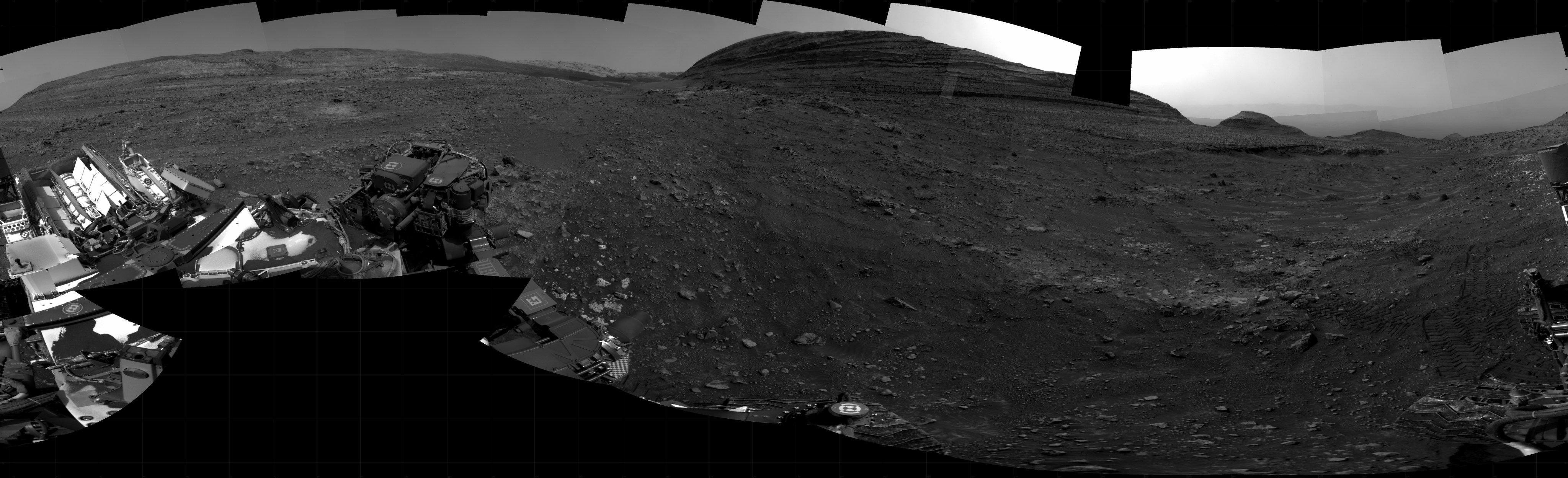 A grayscale panorama of the Martian surface shows a wide field of flat terrain dotted with small rocks, all in dark gray, stretching into the distance where a large butte rises from the ground. A portion of the Curiosity rover is visible in the lower left corner of the image.