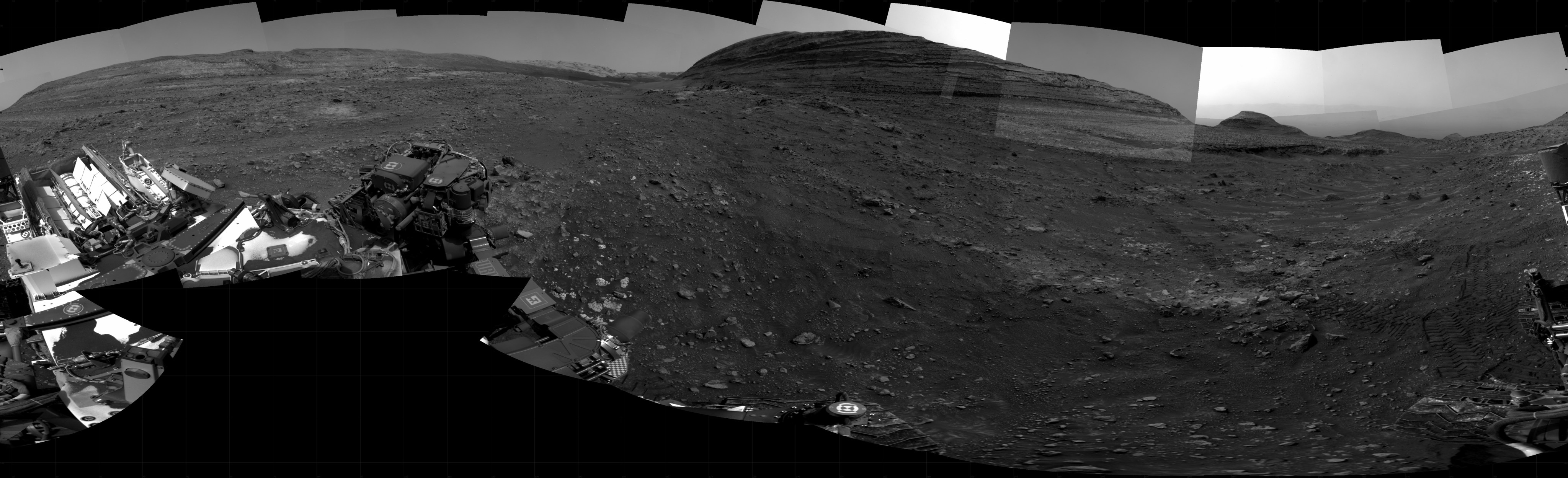 A grayscale panorama of the Martian surface shows a wide field of flat terrain dotted with small rocks, all in dark gray, stretching into the distance where a large butte rises from the ground. A portion of the Curiosity rover is visible in the lower left corner of the image.