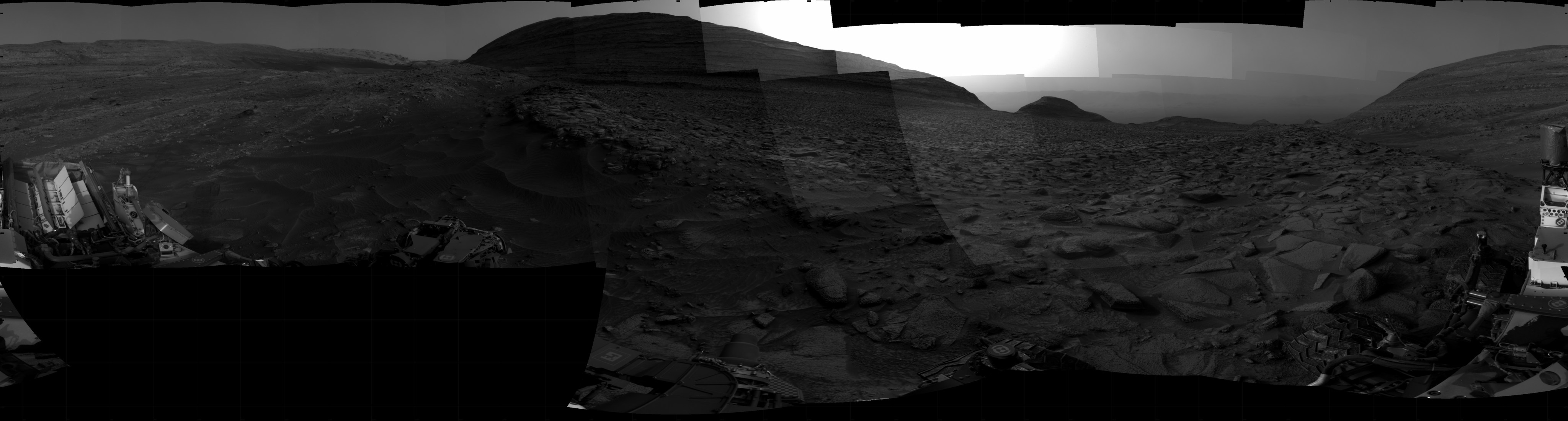 A grayscale panorama of the Martian surface shows night falling over a wide field of flat terrain dotted with small rocks, all in dark gray, stretching into the distance where a large butte rises from the ground. The only bright area of sky remaining is behind the butte. A portion of the Curiosity rover is visible in the lower left and lower right corners of the image.