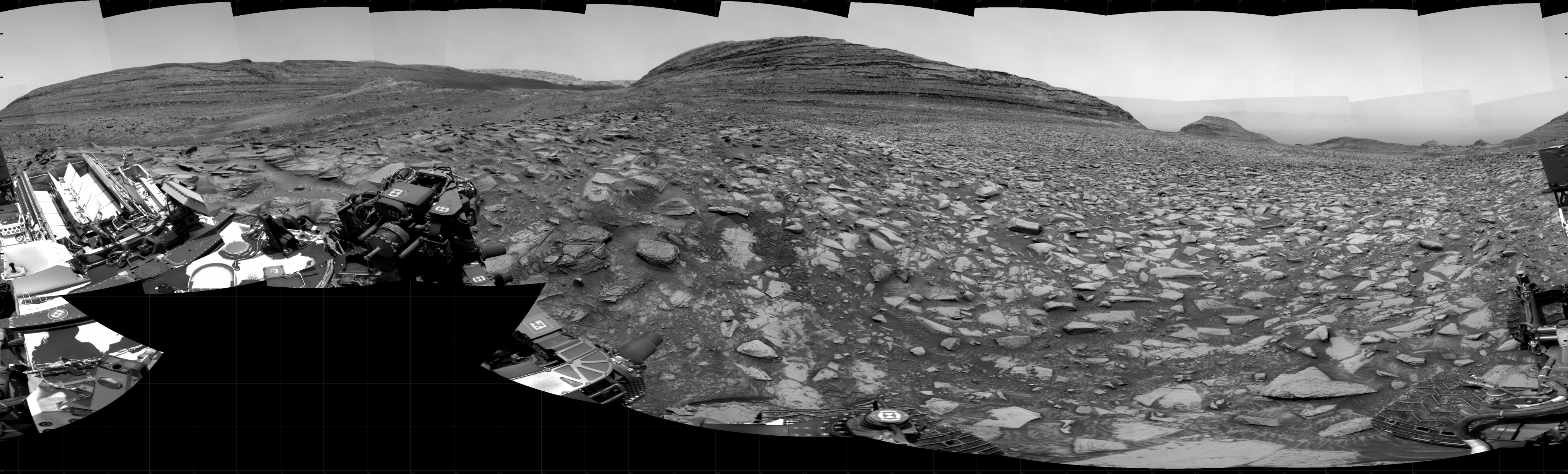 A grayscale panorama of the Martian surface shows a brightly lit, wide field of flat terrain dotted with flat, angular, medium rocks, all in dark gray, stretching into the distance where a large butte rises from the ground. A portion of the Curiosity rover is visible in the lower-left corner and far-right side of the image.