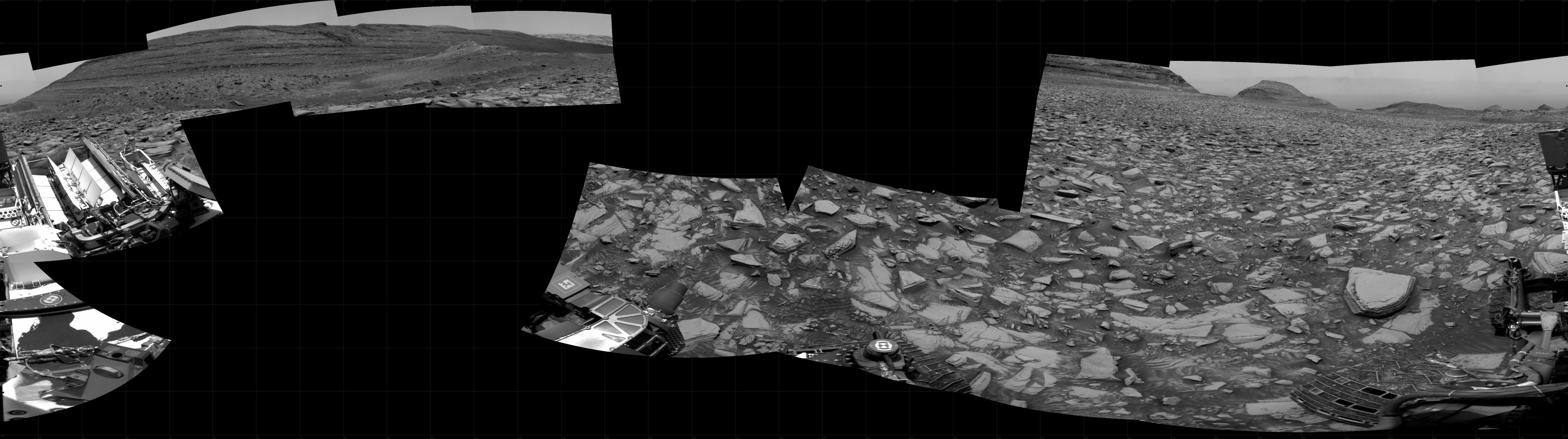 A grayscale panorama of the Martian surface shows a brightly lit, wide field of flat terrain dotted with flat, angular, medium rocks, all in dark gray, stretching into the distance where a series of hills rise from the ground. A portion of the Curiosity rover is visible in the lower-left side of the image.