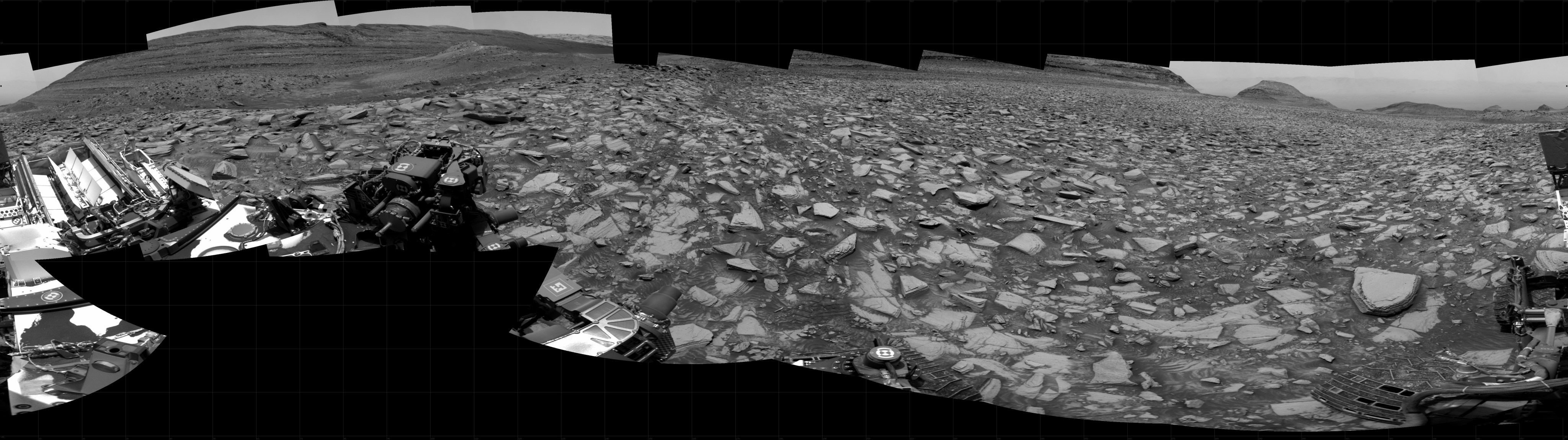 A grayscale panorama of the Martian surface shows a brightly lit, wide field of flat terrain dotted with flat, angular, medium rocks, all in dark gray, stretching into the distance where a series of hills rise from the ground. A portion of the Curiosity rover is visible in the lower-left side of the image.