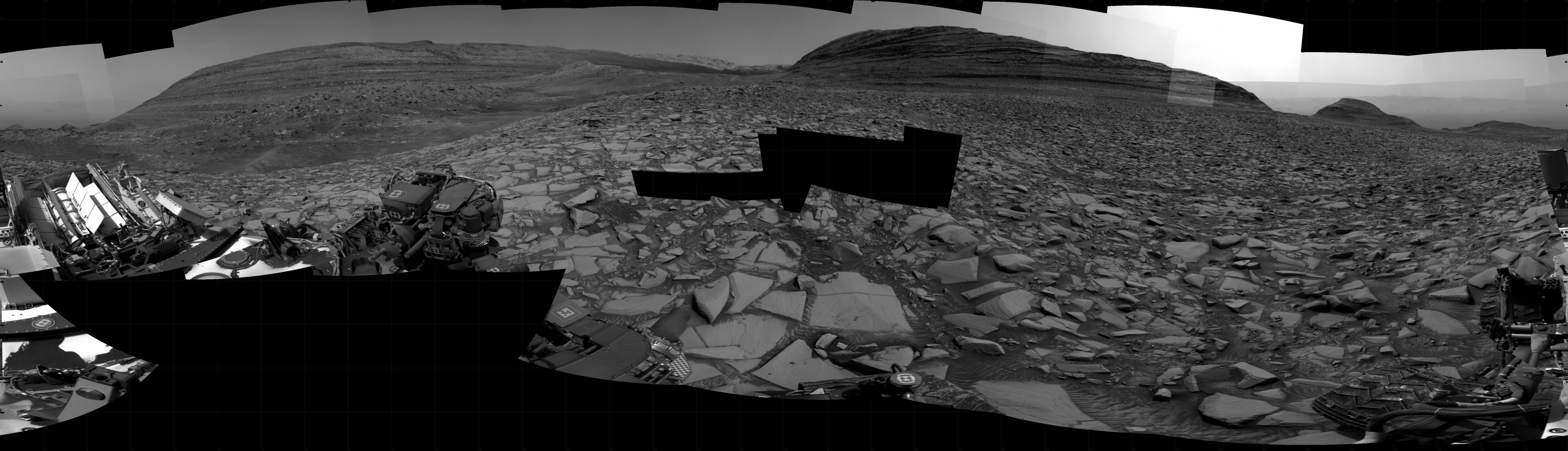 A grayscale panorama of the Martian surface shows a wide field of flat terrain dotted with flat, angular, medium-sized rocks, all in dark gray, stretching into the distance where a series of hills rise from the ground. A portion of the Curiosity rover is visible in the left and right sides of the image.