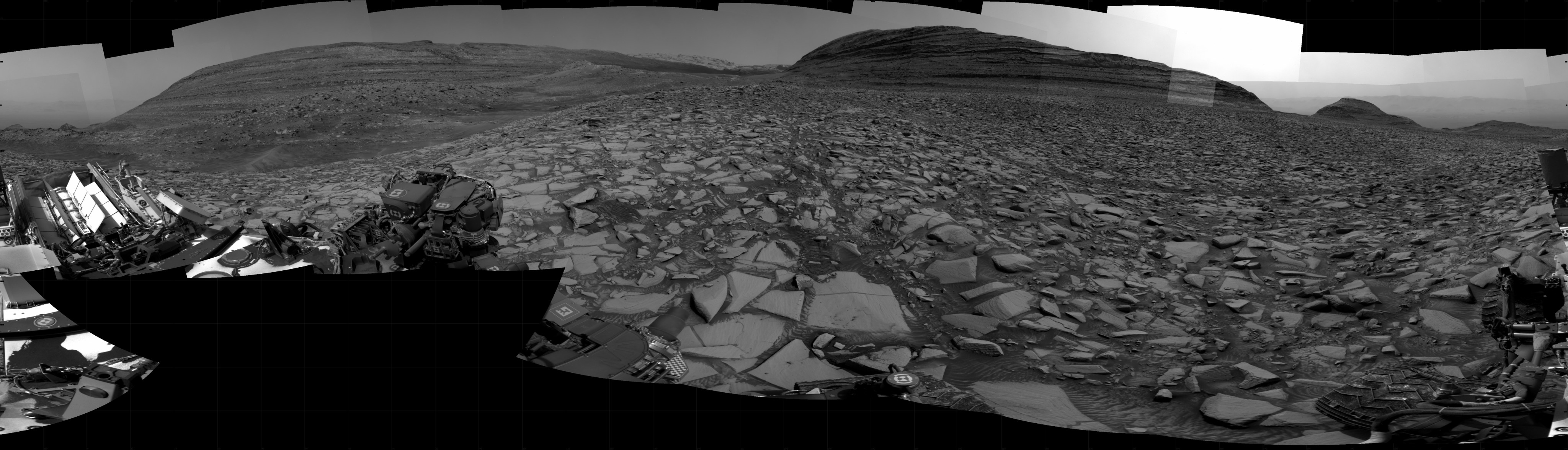 A grayscale panorama of the Martian surface shows a wide field of flat terrain dotted with flat, angular, medium-sized rocks, all in dark gray, stretching into the distance where a series of hills rise from the ground. A portion of the Curiosity rover is visible at the left and right sides of the image.