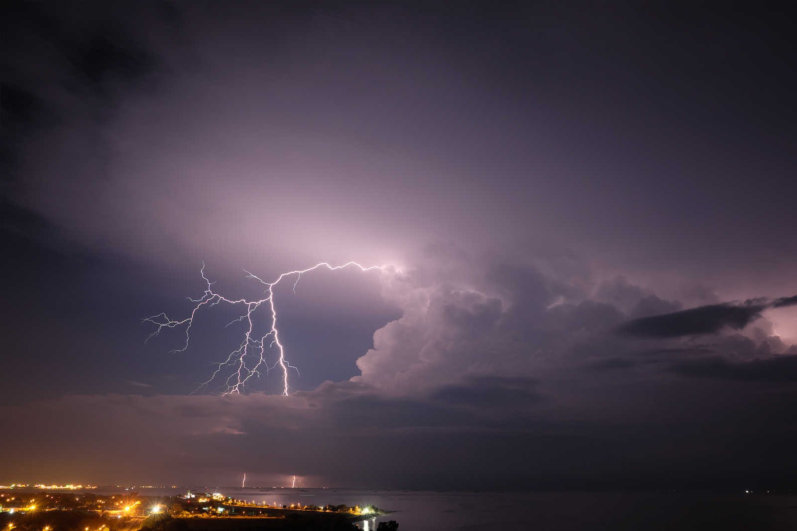 Two lightning strikes are visible in the distance at night with city lights along the left of the image along the coastline of the bay that occupies the lower right and curves into the landscape on the lower left. The majority of the image is occupied by dark gray cloudcover.