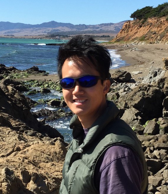 Young man with blue sunglasses and dark hair stands on a rocky beach. Shallow desert mountains stretch out in the background.